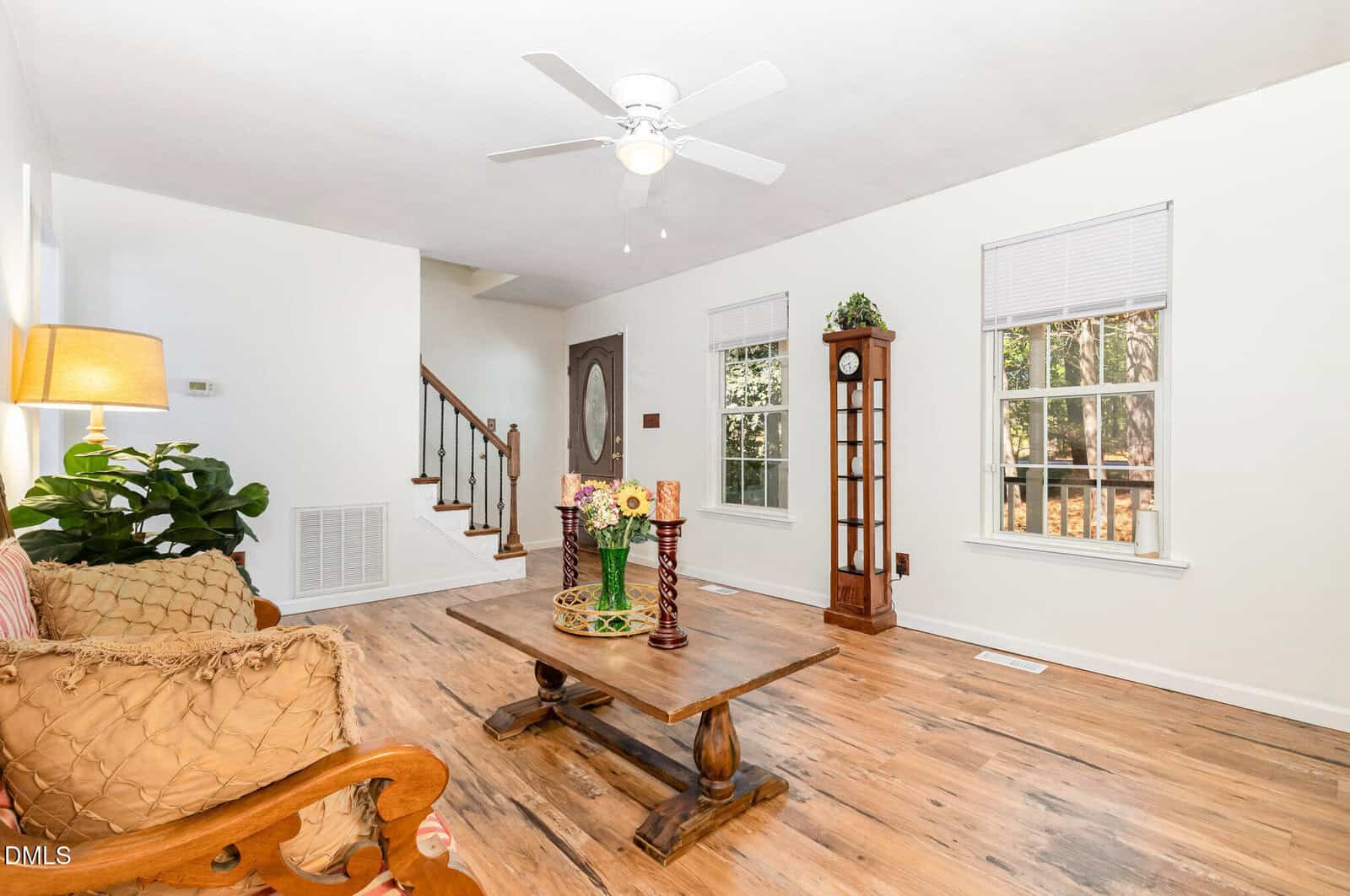 Bright living room with white walls, wooden floors, a wooden coffee table with flowers, a cushioned chair, a tall grandfather clock, potted plants, and large windows letting in natural light. Ceiling fan overhead.
