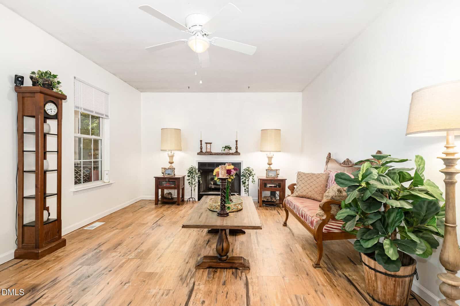 Bright living room with wood floor, a ceiling fan, vintage sofa, coffee table, tall clock, two side tables with lamps, fireplace, potted plants, and a floor lamp. White walls and large window on the left.