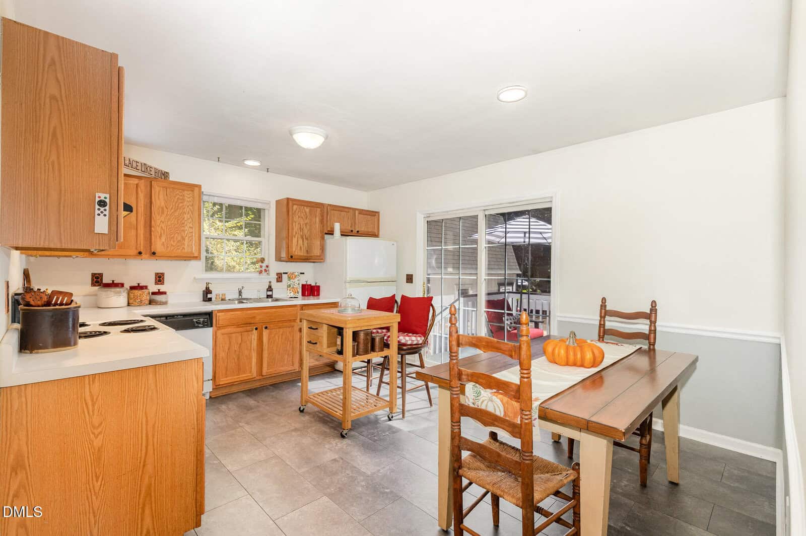 A kitchen with wooden cabinets, white appliances, a small center island, and a dining table set with three chairs and pumpkins. A sliding glass door leads to an outdoor patio. Light gray tile flooring throughout.