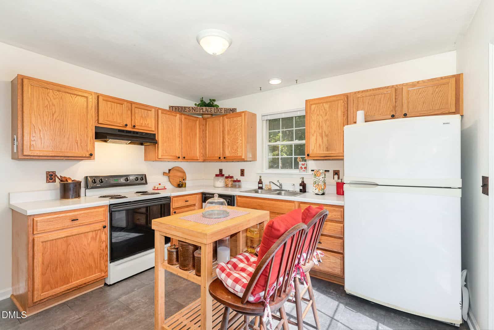 A kitchen with wooden cabinets, white countertops, a white refrigerator, electric stove, sink under a window, and a small wooden island with two stools. Kitchen items and decor are visible on the counters and shelves.