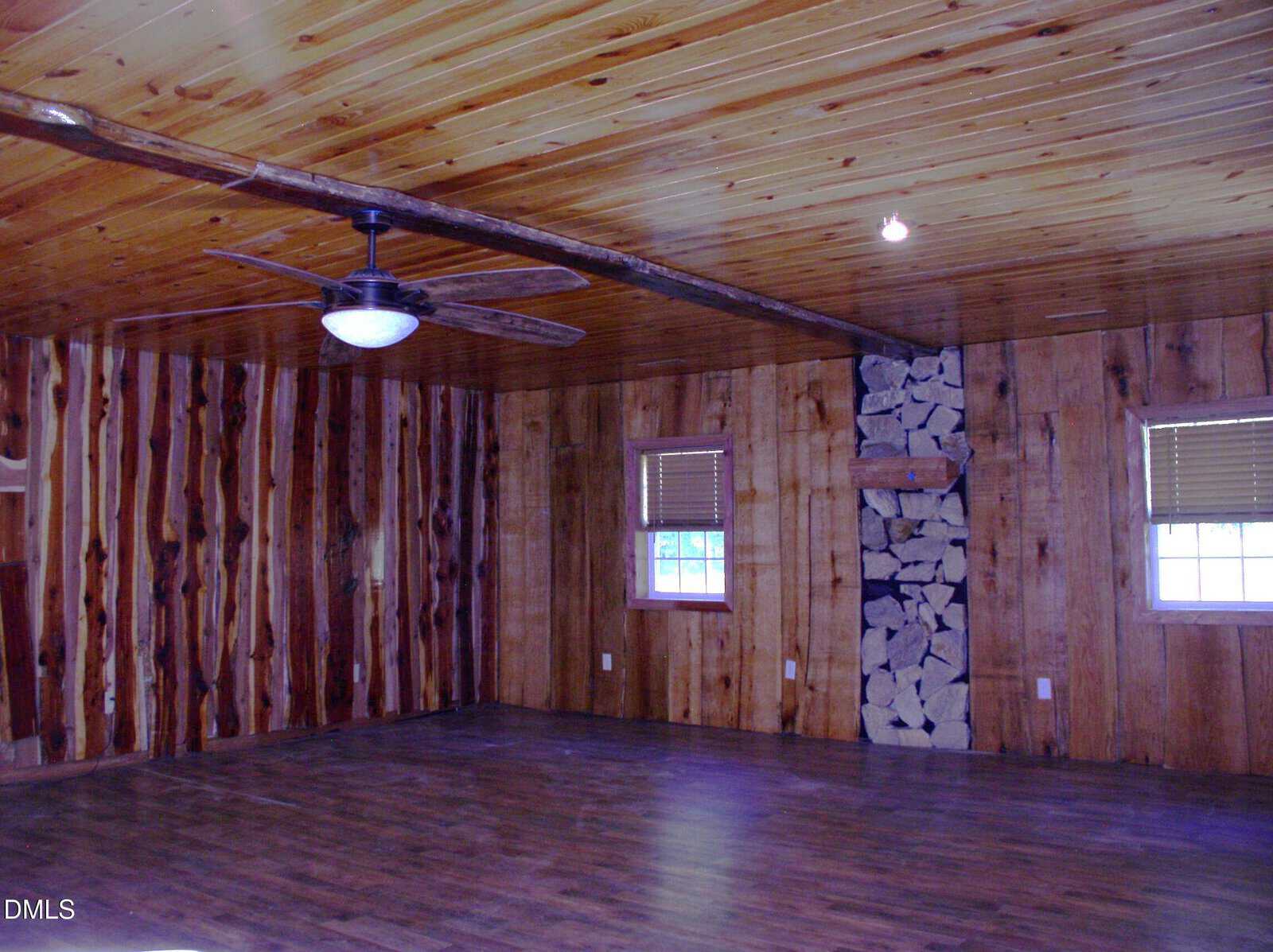 A large empty room with wood-paneled walls and ceiling, hardwood floors, two windows with blinds, a ceiling fan with a light, and a vertical stack of firewood built into one wall.