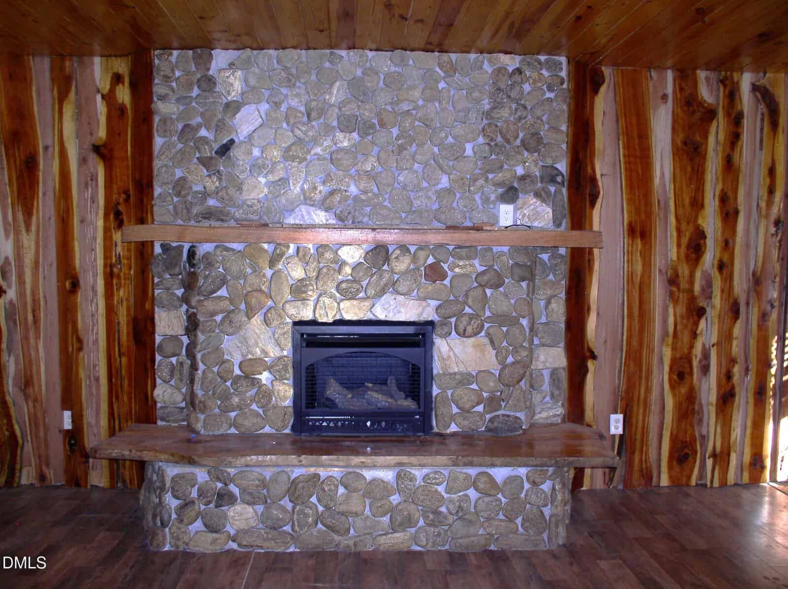 Stone fireplace with wood mantel and bench, set against a wall and ceiling covered in natural wood planks. The floor is dark wood, and electrical outlets are visible on both sides of the fireplace.