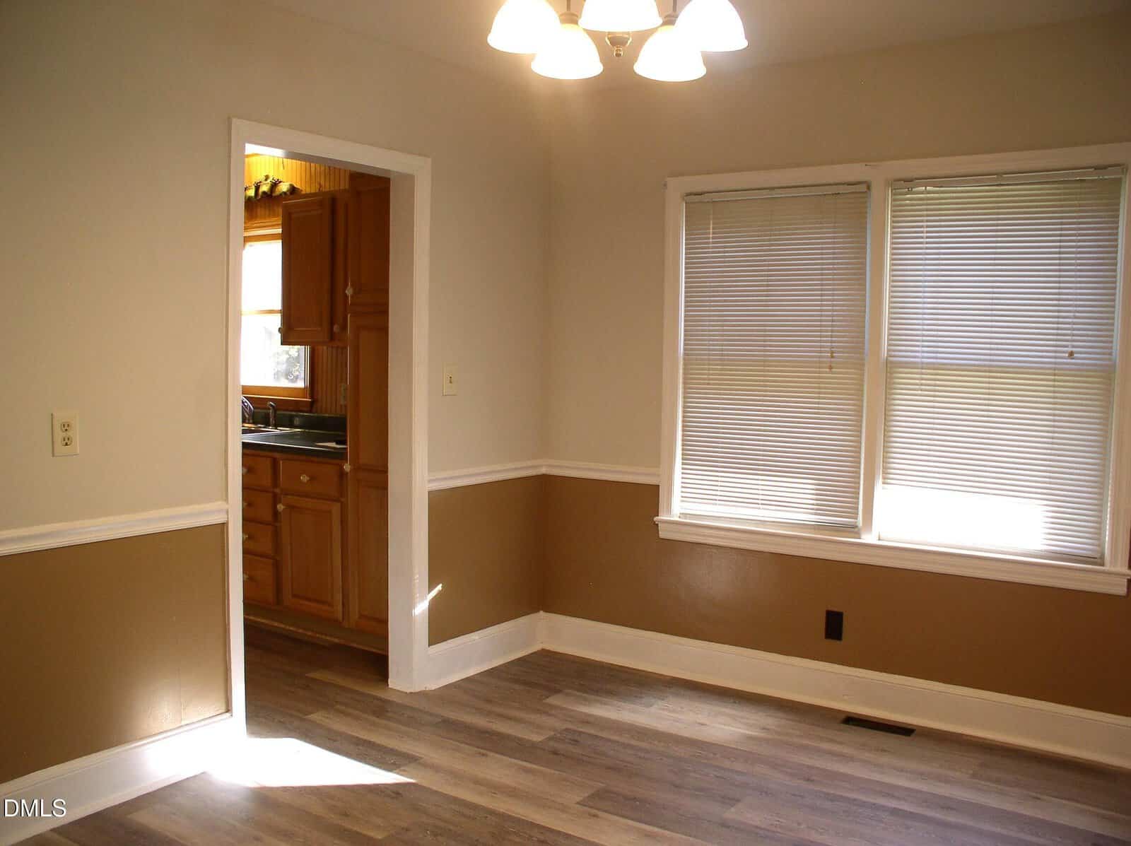 A dining room with tan and white walls, wood flooring, and two windows with blinds. An open doorway leads to a kitchen with wooden cabinets and a sink visible. Ceiling light fixture is on.