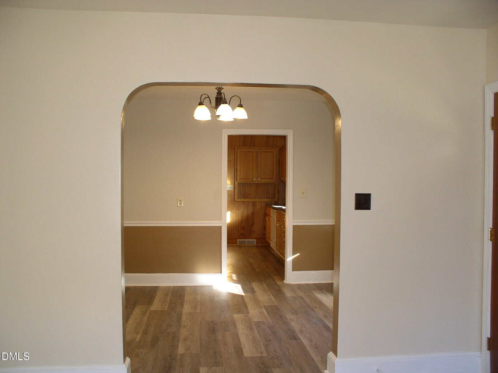 View through an arched doorway into a dining room with a ceiling light fixture, beige walls with a brown lower half, and wood flooring, leading into a kitchen with wood cabinets.