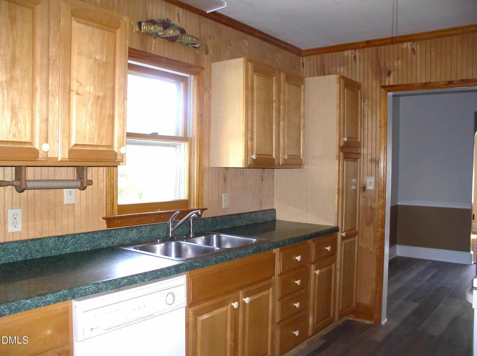 A kitchen with wood cabinets, green countertops, a double sink under a window, a dishwasher, and wood-paneled walls. The floor is dark with a visible adjoining room in the background.