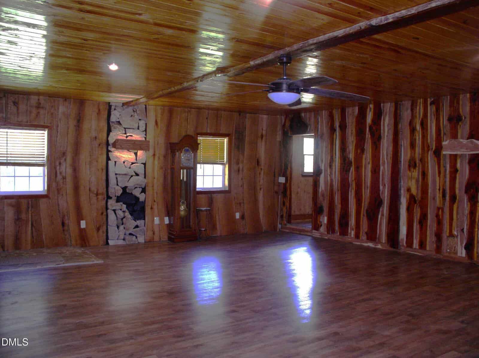 A spacious room features wood-paneled walls and a polished wood ceiling with exposed beams. There are several windows, a ceiling fan with lights, a stone fireplace, and a grandfather clock. The floor appears to be laminate wood.