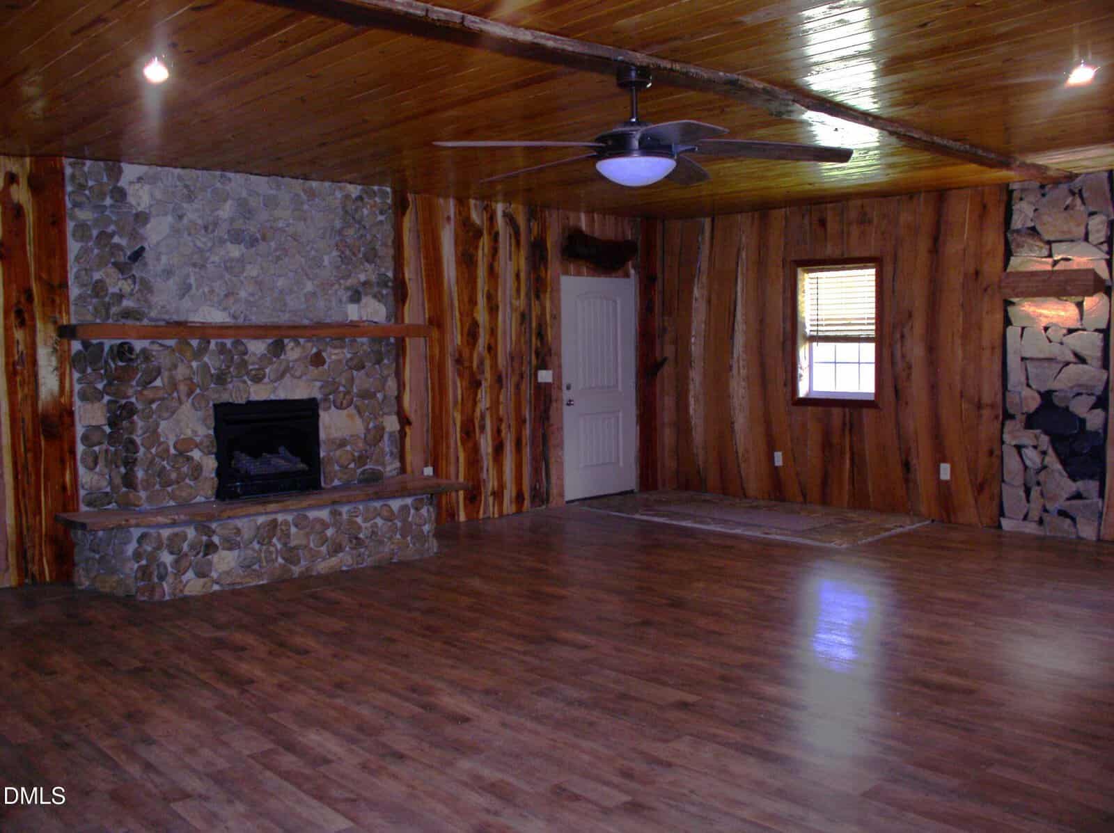 A spacious room with wood-paneled walls and ceiling, stone fireplace, ceiling fan, single door, a window with blinds, and laminate wood flooring. Stonework decor is also featured on the right wall.