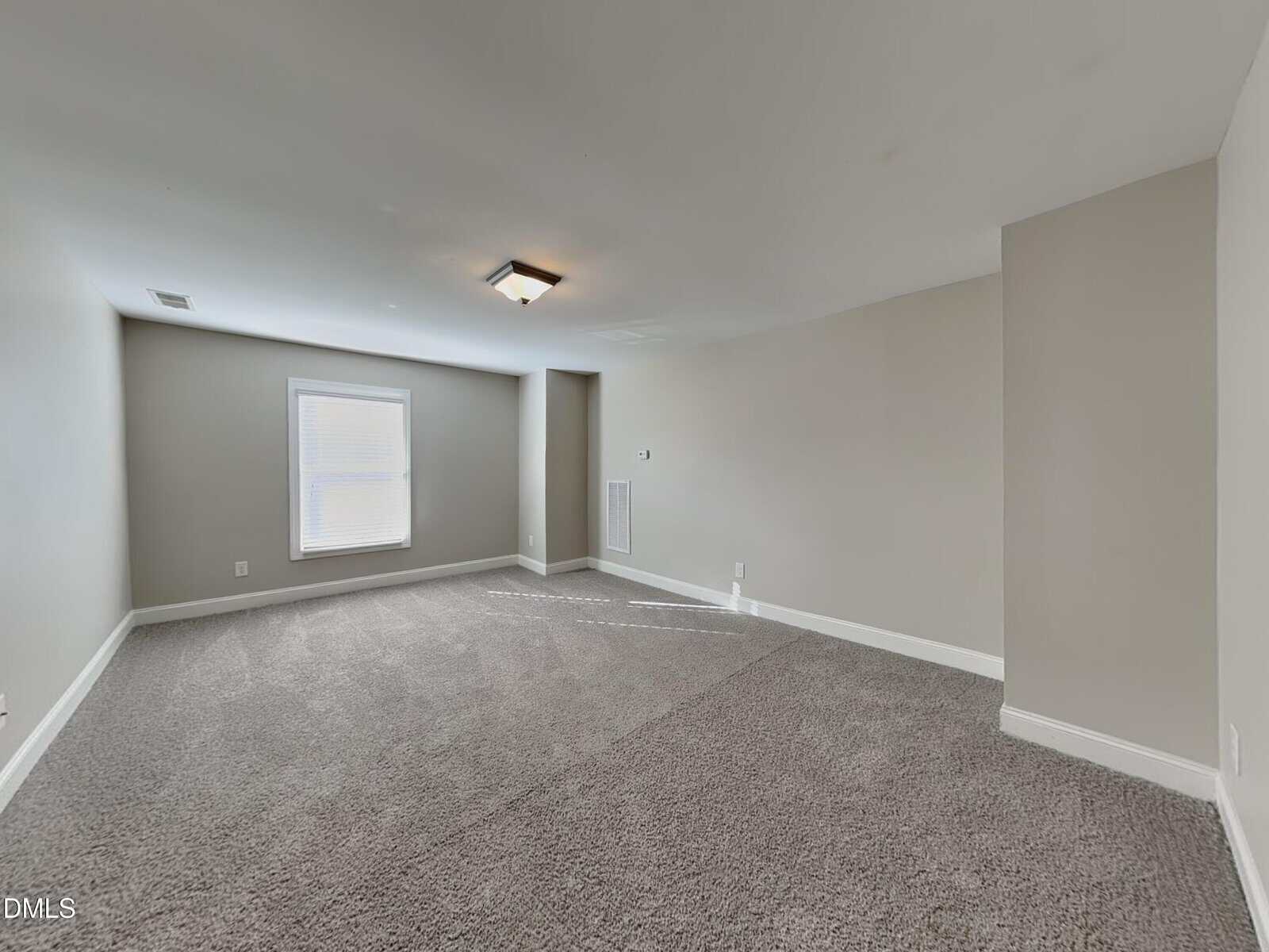 A carpeted, empty room with beige walls, a ceiling light fixture, one window with blinds, and white baseboards. The room appears clean and well-lit with natural light coming from the window.