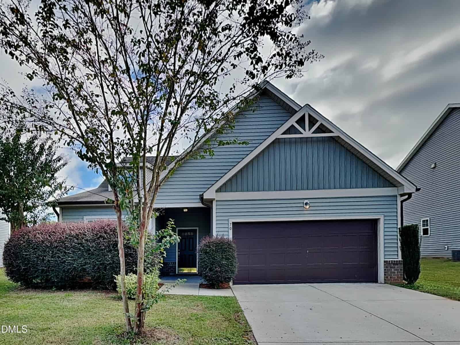 A one-story gray house at 70 Leaf Spring Way, Youngsville, with a dark two-car garage, gable roof, and white trim. A tree and trimmed bushes line the front yard, and a concrete driveway leads to the garage under a partly cloudy sky.