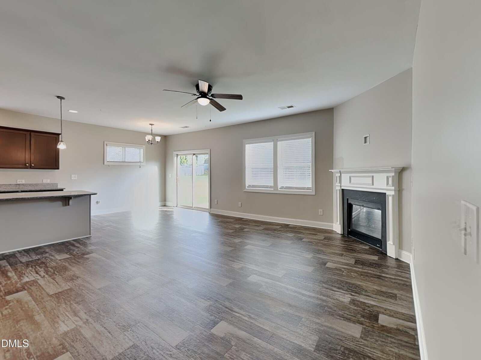 Open-concept living area with wood-style floors, a ceiling fan, fireplace, large windows, and a sliding glass door. Kitchen with dark cabinets and a breakfast bar is visible to the left. Neutral-colored walls throughout.
