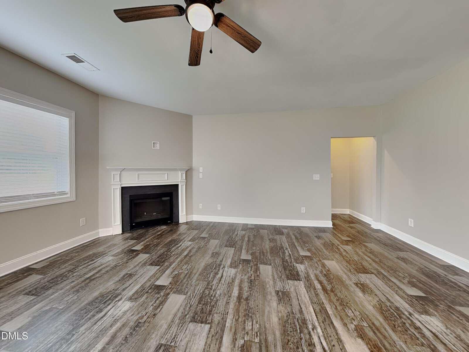 A spacious, empty room with wood-look flooring, a ceiling fan with lights, a white fireplace in the corner, gray walls, and a window on the left. There is a doorway leading to another area.