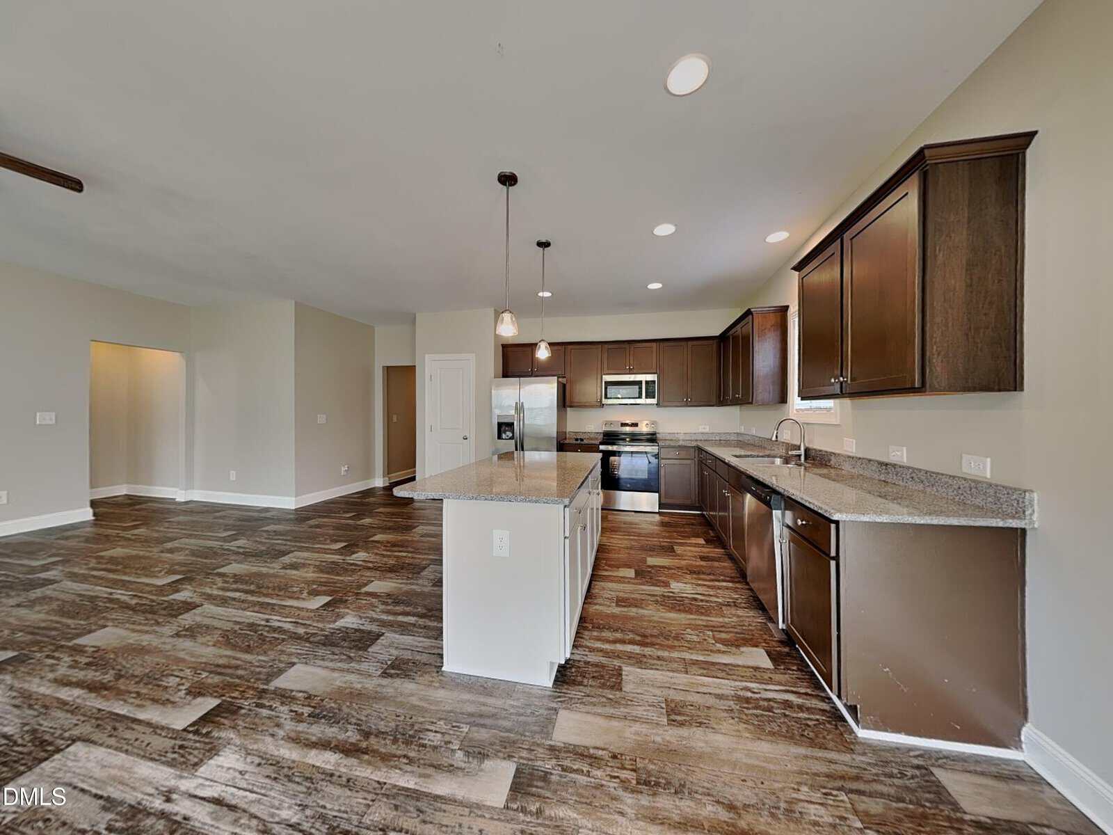 Spacious modern kitchen with dark wood cabinets, stainless steel appliances, granite countertops, island with sink, pendant lighting, and wood-look flooring extending into an open living area.