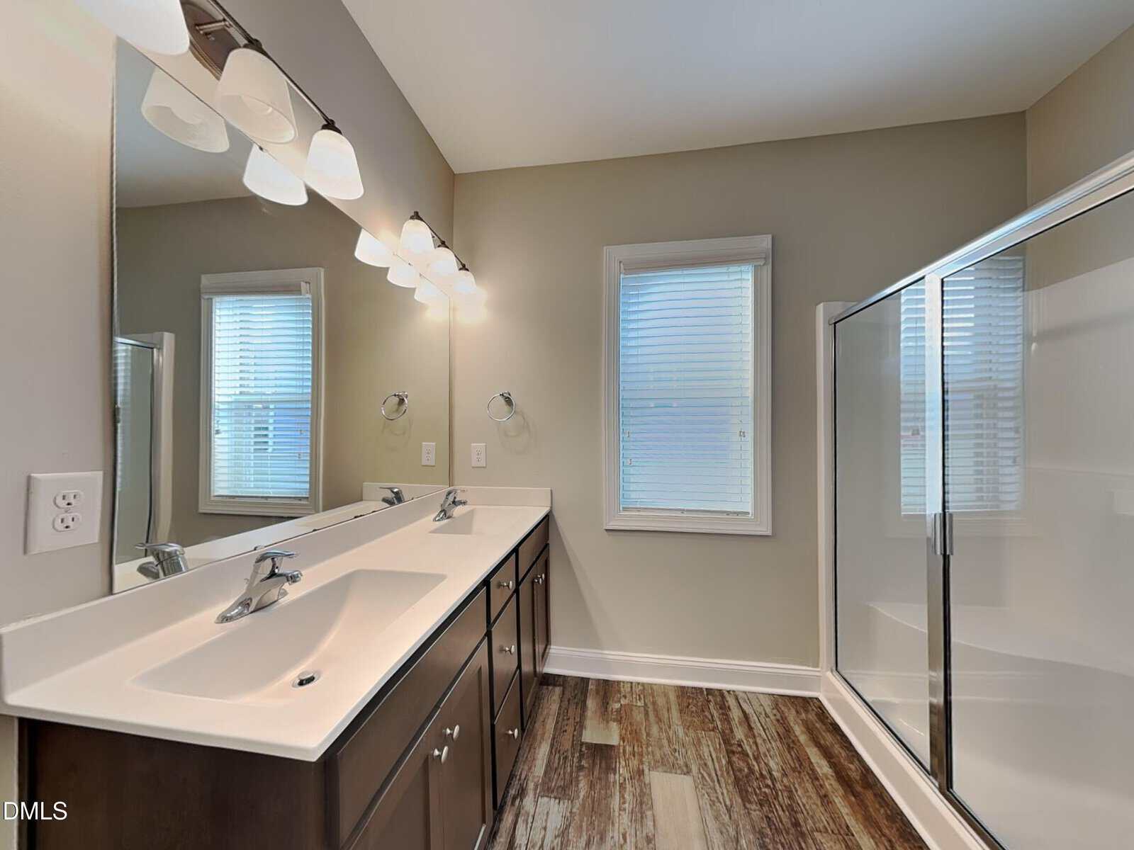 A bathroom with a double sink vanity, a wide mirror with five lights above, a window with blinds, and a glass-enclosed shower. The floor has a wood-look design, and the walls are light-colored.