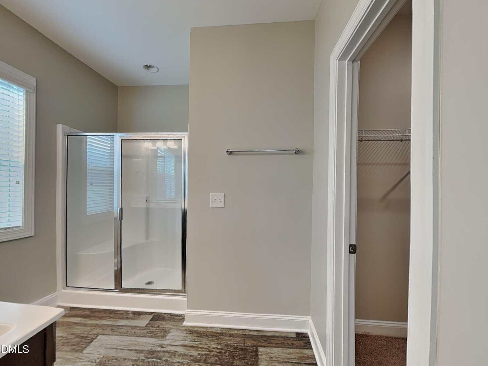 A bathroom with a glass-enclosed shower, a towel bar on the wall, a window to the left, and an open closet with shelves and carpeted flooring to the right. The floor is wood-patterned.