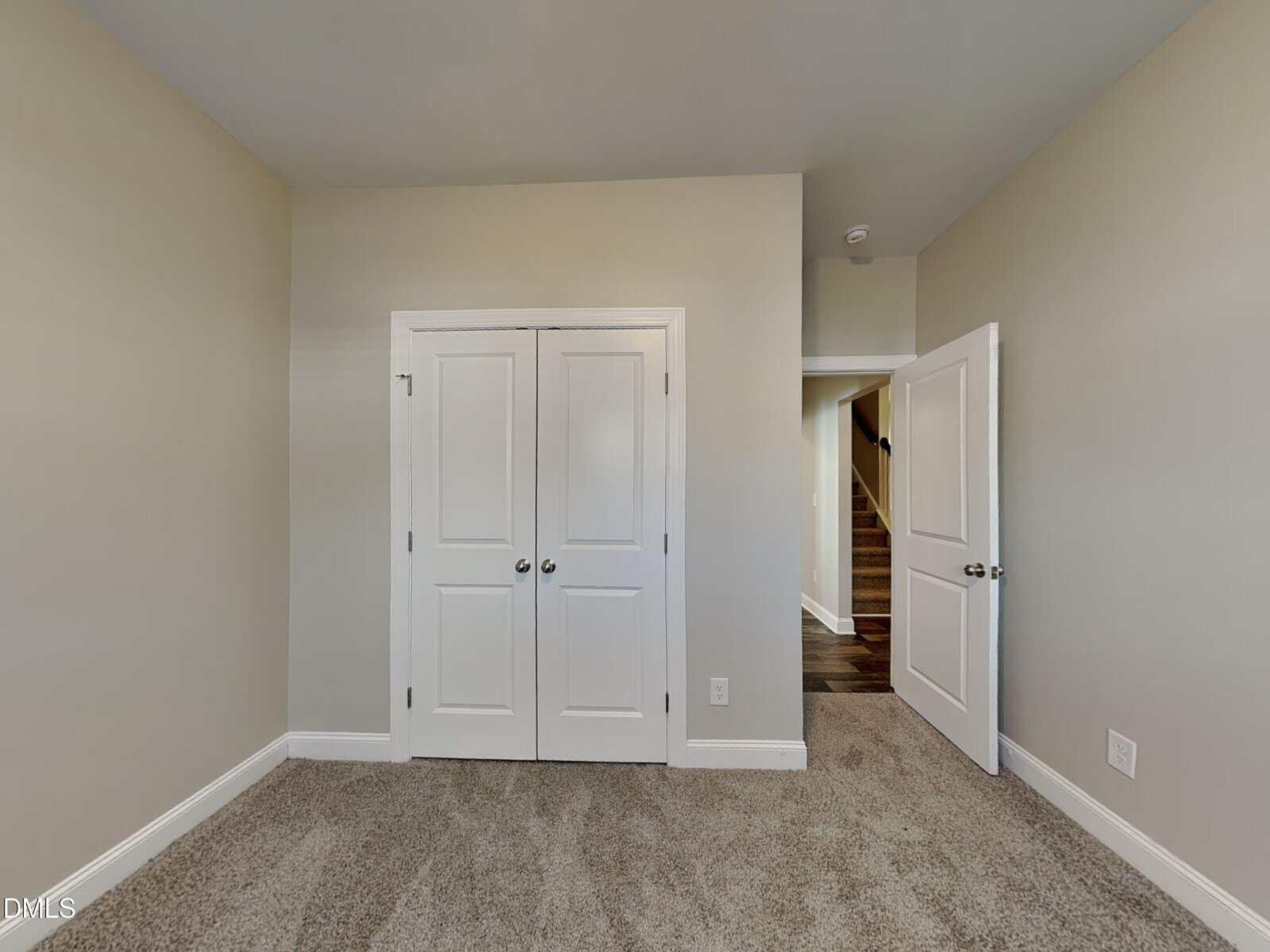 A small empty room with beige walls, white double closet doors, carpeted floor, and an open white door leading to a hallway with stairs.