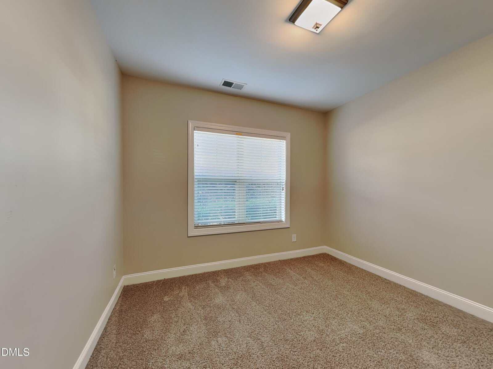 Empty room with beige walls, a carpeted floor, a large window with blinds, white baseboards, and a ceiling light fixture.