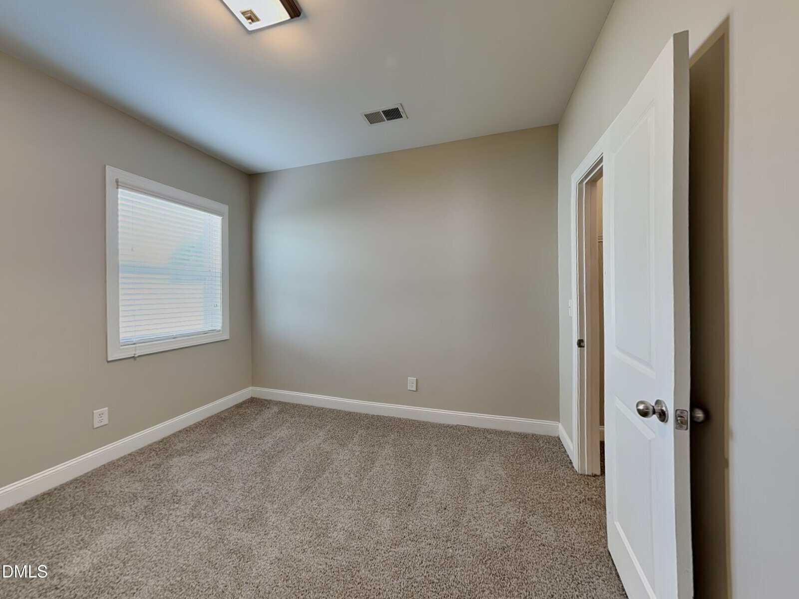 Empty bedroom with beige walls, a single window with blinds, gray carpet, an open white door, and a ceiling light. The room is well-lit and minimalistic, with vents on the ceiling and baseboard trim.