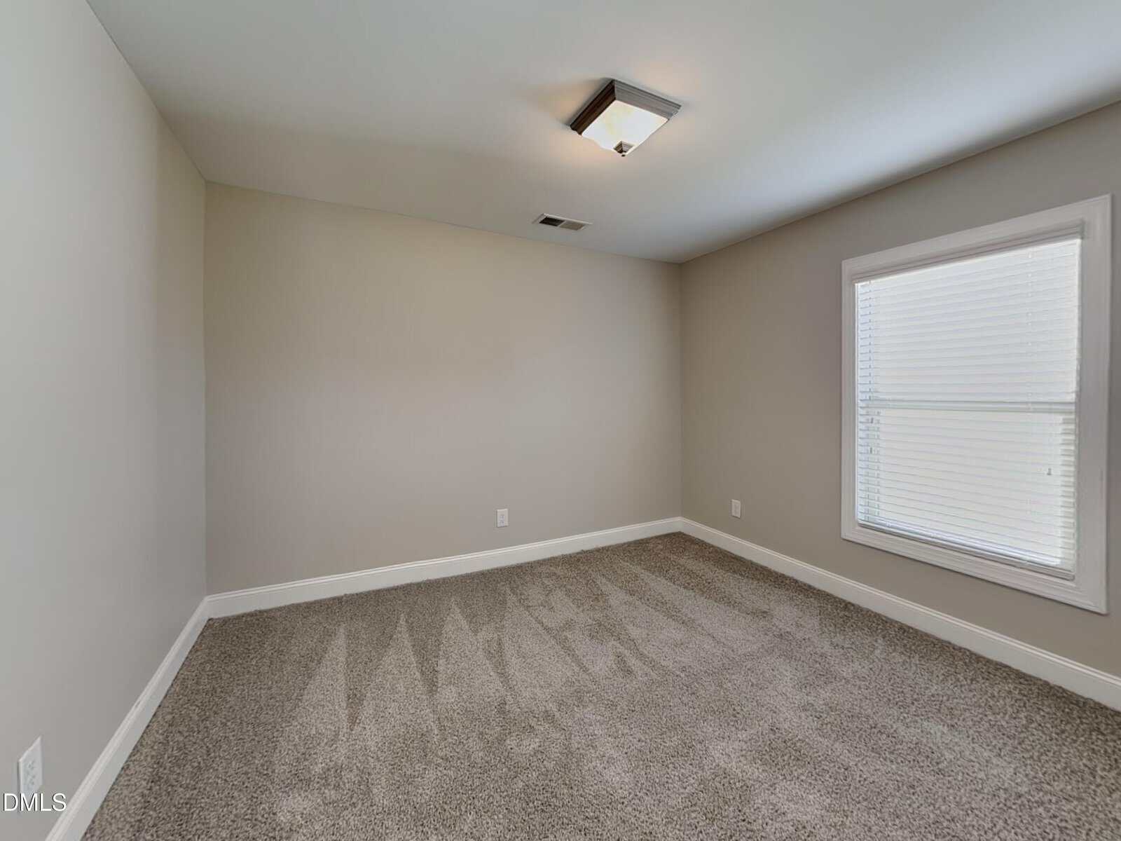 An empty room with beige walls, gray carpet, a ceiling light, a single window with blinds, and white trim along the floor and window.
