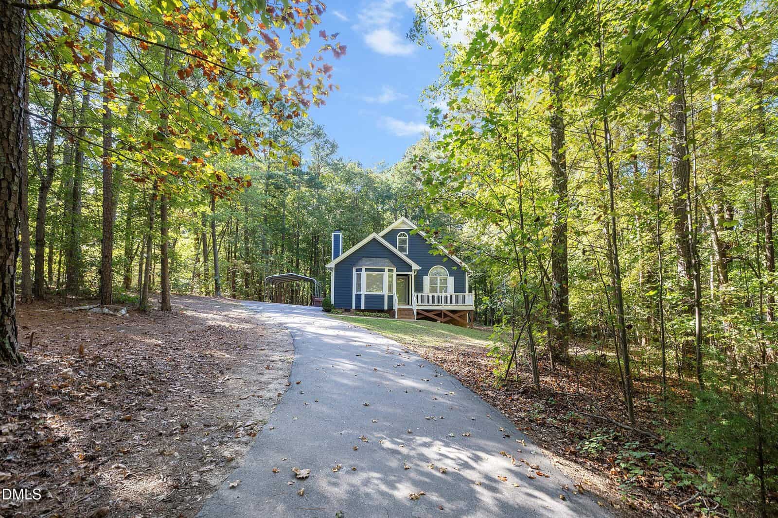 A paved driveway leads through a wooded area to a blue house with white trim, front porch, and dormer windows, surrounded by tall trees and greenery under a clear sky.