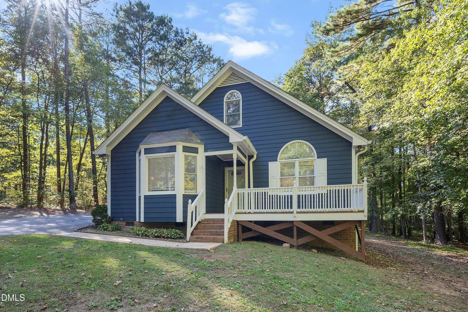 A blue house with white trim at 1775 Sid Mitchell Road features a front porch, bay window, and sloped roof. Surrounded by trees in peaceful Youngsville, the home sits on a grassy yard with a driveway leading to the left. Steps lead up to the porch and front door.