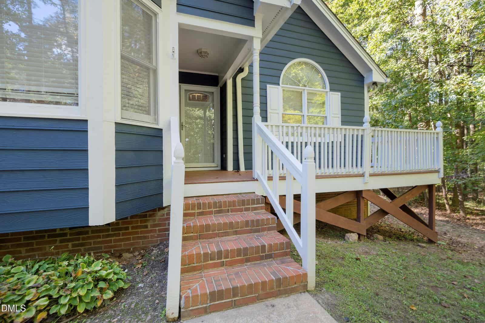 Front entrance of a blue house with white trim, brick steps, a small porch with railing, and a glass front door. The house is surrounded by trees and partial shade.