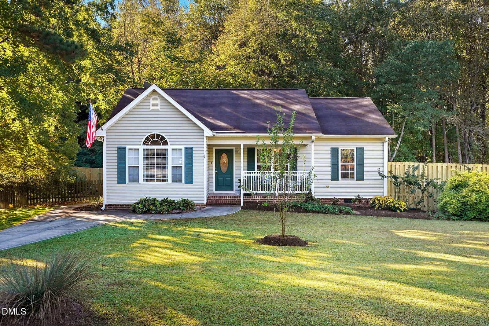 Charming single-story beige house at 55 Meadowfield Lane, Franklinton, featuring teal shutters, a welcoming front porch, and an American flag. The home showcases a sloped roof, small front garden, young tree, and driveway—all set against the tranquil backdrop of a wooded area.