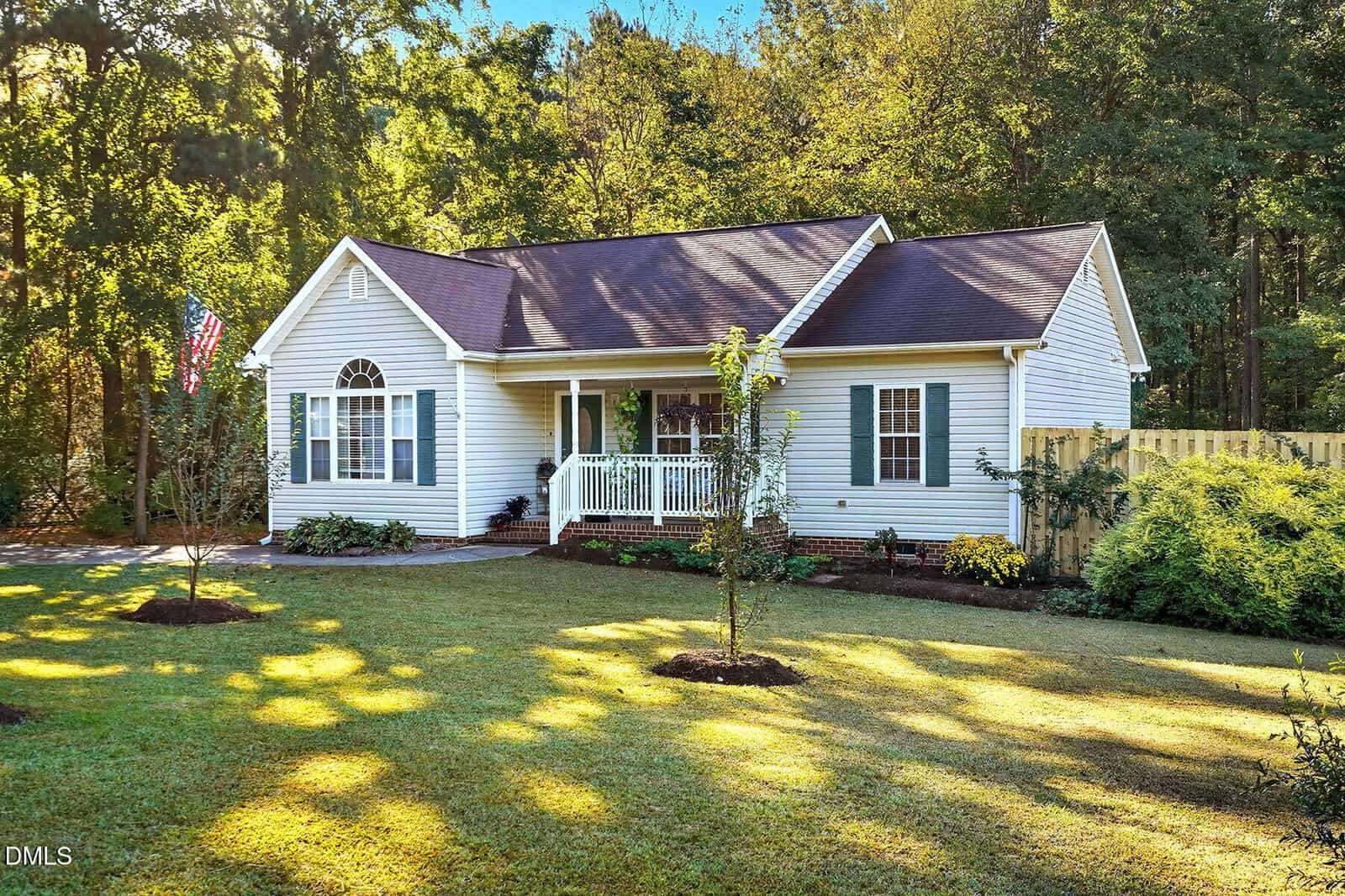 Single-story white house with dark roof, front porch, and shutters. The home is surrounded by a green lawn, young trees, bushes, and a wooden fence. Tall trees are visible in the background. The weather is sunny and clear.