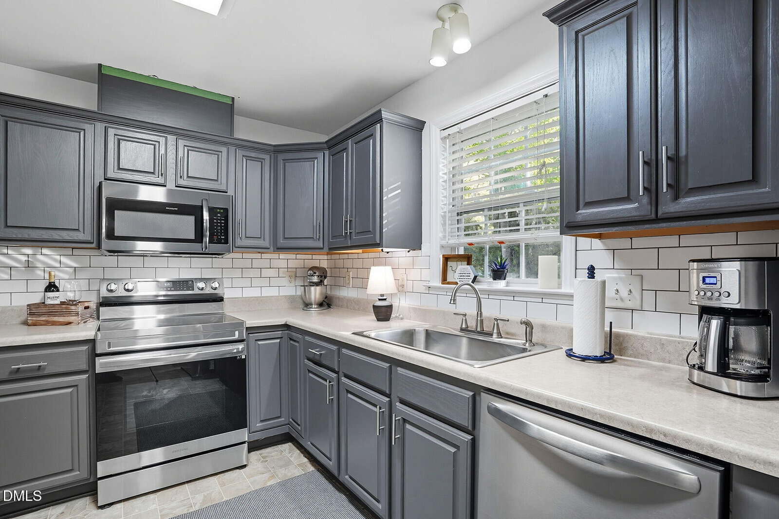 Modern kitchen with gray cabinets, stainless steel appliances, white subway tile backsplash, coffee maker, and a window above the sink with blinds and plants on the sill. Countertops are light-colored, and lighting is overhead.
