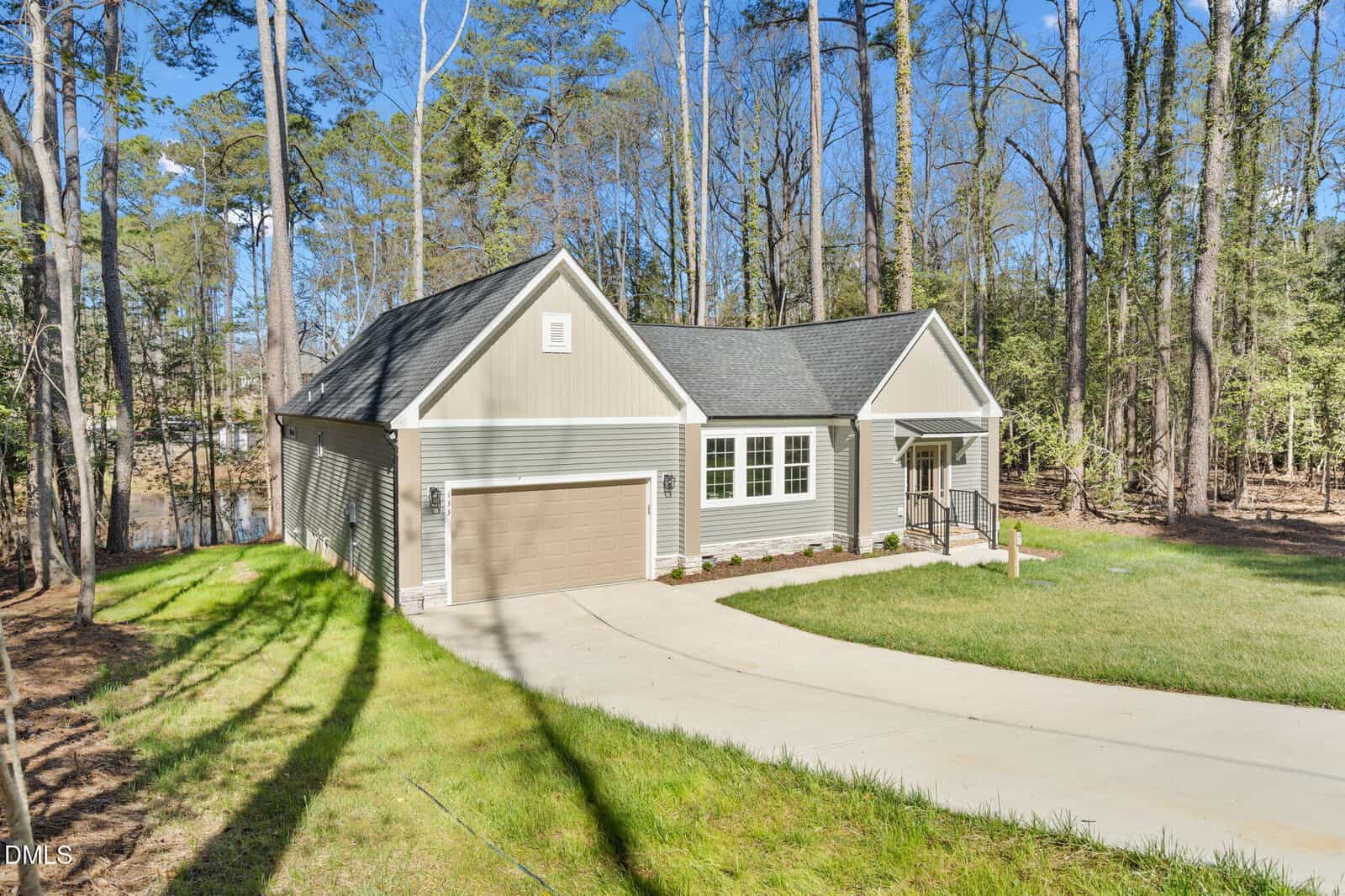 A light gray, single-story house with a two-car garage, surrounded by tall trees and a well-kept lawn, sits at the end of a curved concrete driveway under a clear blue sky.