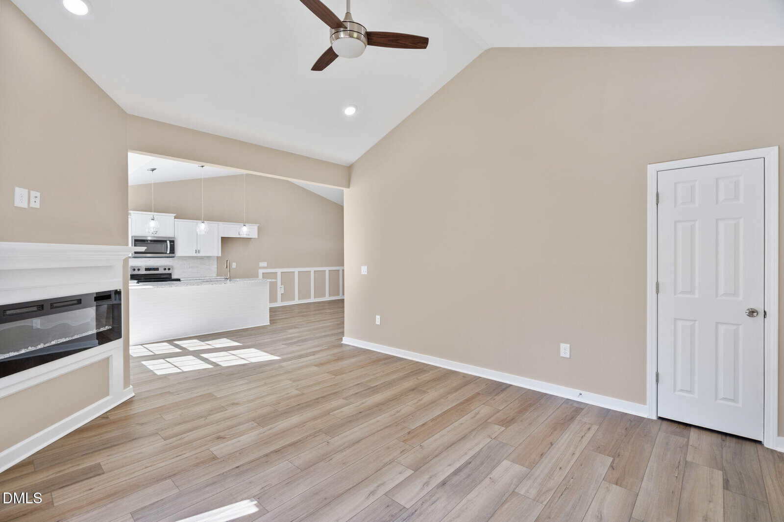 Bright, modern open-concept living space with light wood floors, beige walls, vaulted ceiling, ceiling fan, electric fireplace, and a view into a white kitchen with stainless steel appliances and pendant lighting.