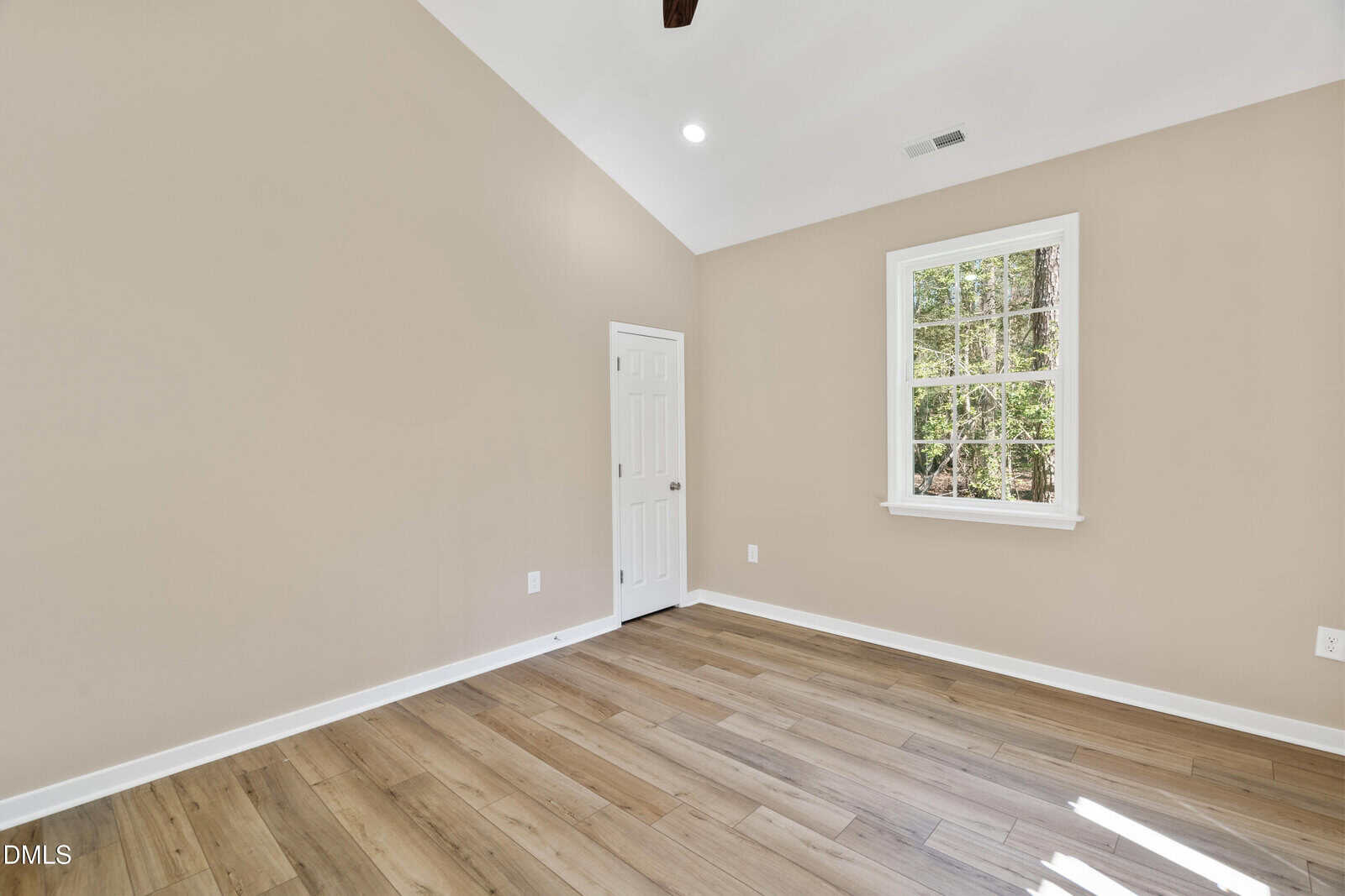 A bright, empty room with beige walls, wood flooring, a white door, and a window letting in natural light. The ceiling is vaulted with recessed lighting and a beam.