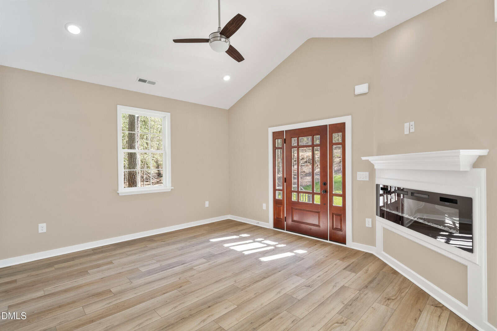 A bright, empty living room with light wood floors, beige walls, vaulted ceiling, ceiling fan, white trim, a window, a modern wall-mounted fireplace, and a large wooden front door with glass panels.