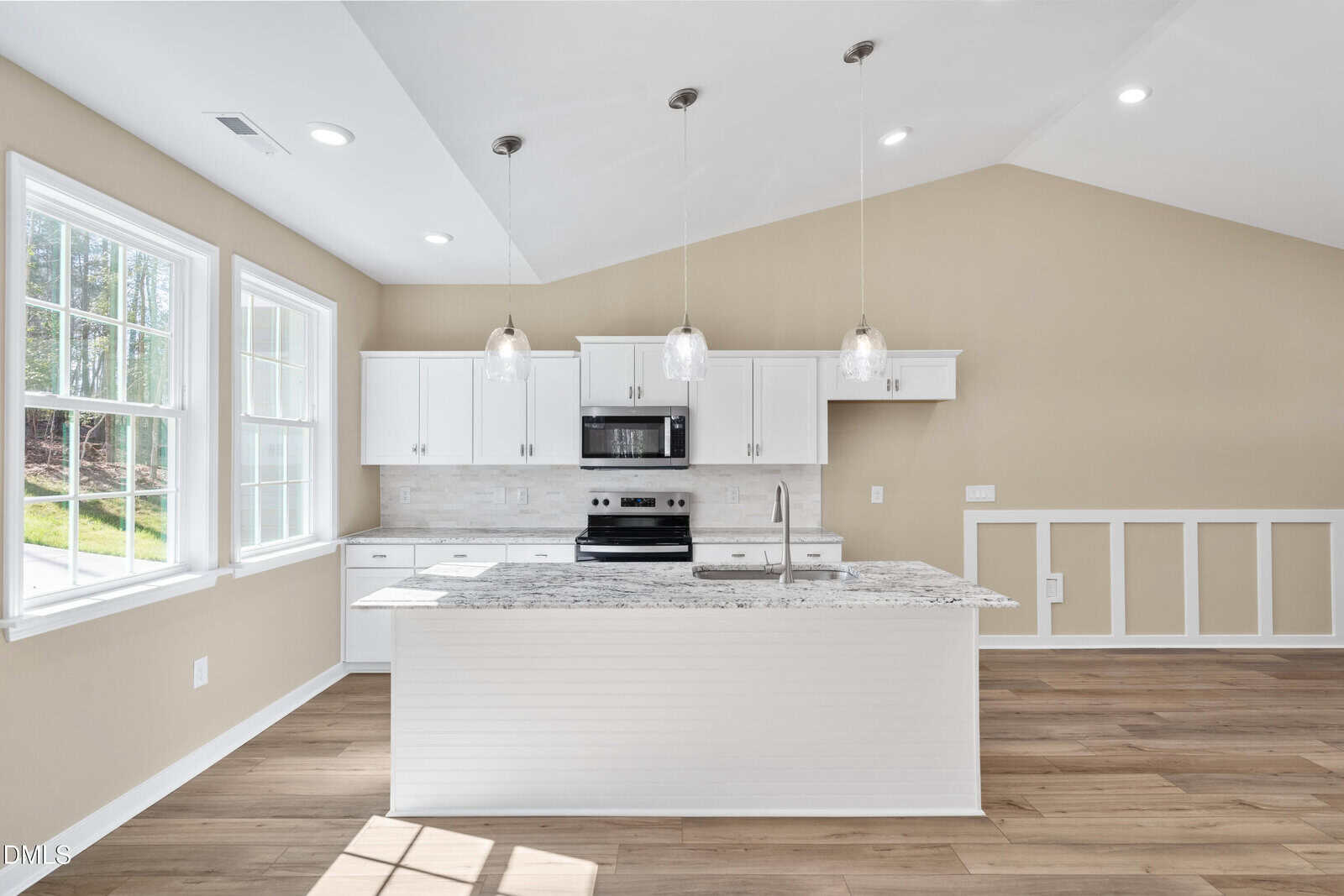 Bright modern kitchen with white cabinets, granite countertops, a central island with sink, stainless steel appliances, pendant lighting, wood flooring, and large windows letting in natural light.
