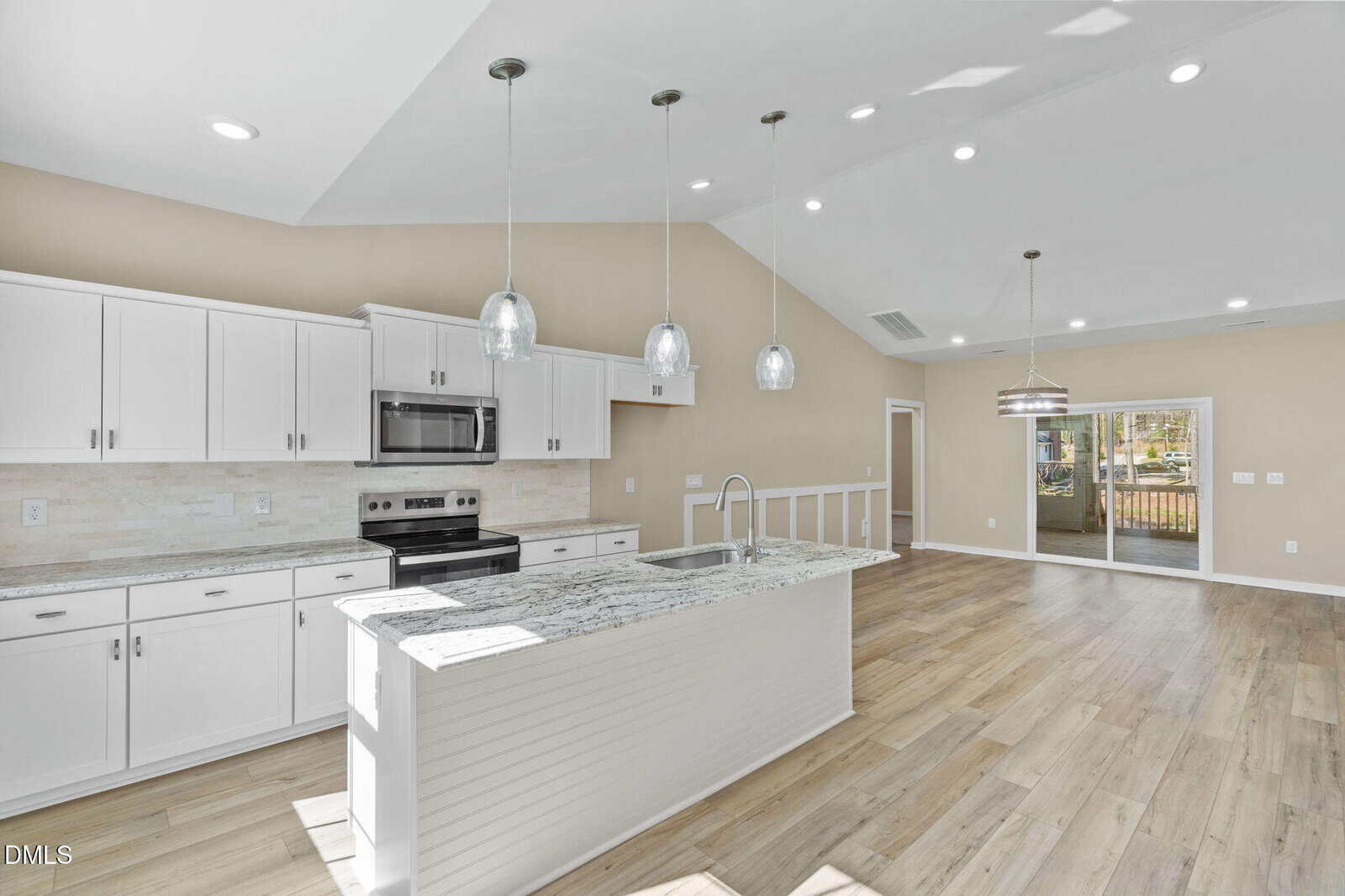 Bright, modern kitchen with white cabinets, stainless steel appliances, granite countertops, pendant lighting over an island, and an open floor plan leading to a dining area with large sliding glass doors.
