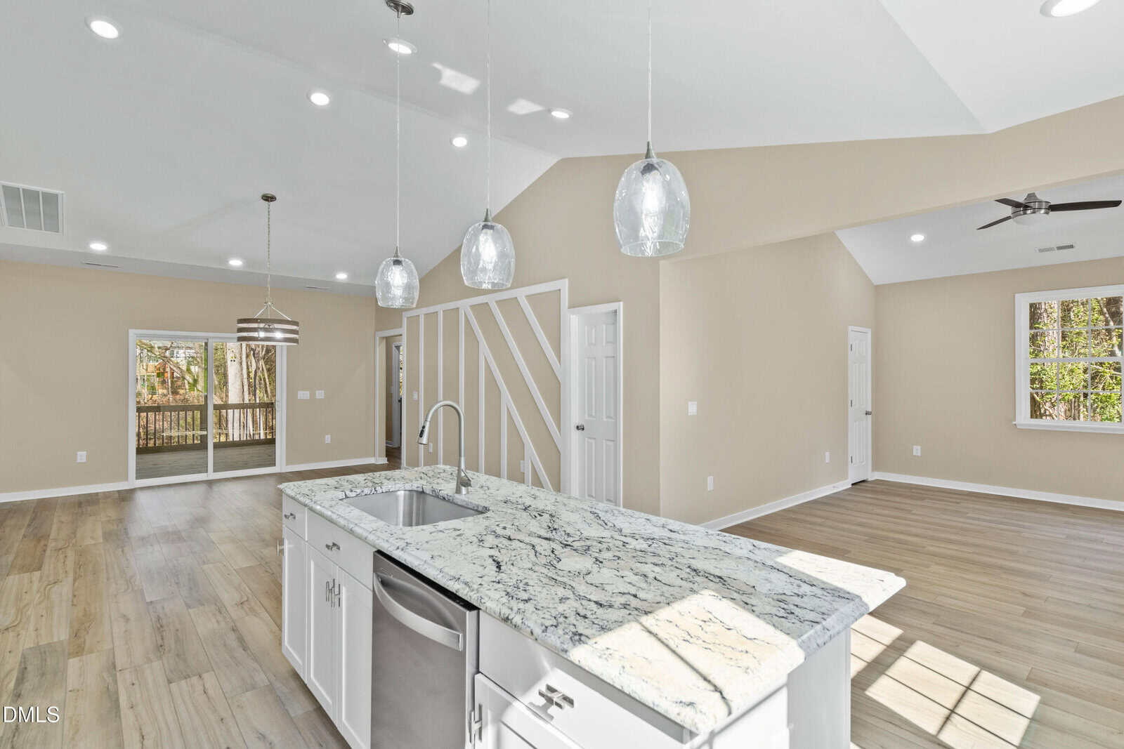Modern open-concept kitchen and living area with light wood floors, white cabinetry, granite countertops, pendant lights, and large windows letting in natural light. Dining area with sliding glass doors to a deck.