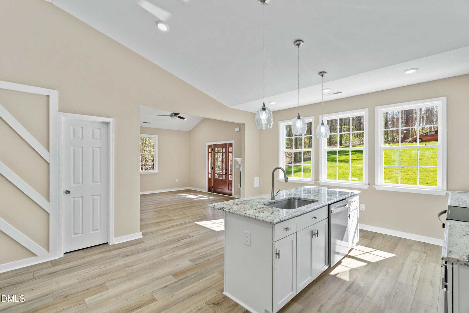 Modern open-concept kitchen and living area with large windows, white cabinets, marble countertops, pendant lights, light wood flooring, and a view of a grassy yard outside.