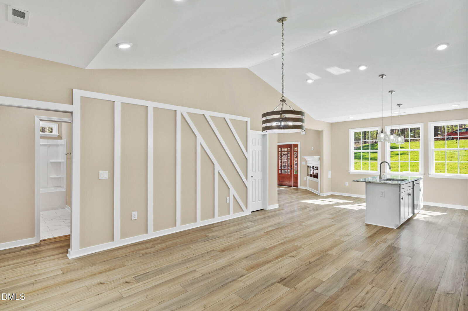 Open-concept living area with light wood floors, beige walls, white trim, a kitchen island, pendant lights, large windows, and a modern accent wall with geometric trim. A doorway leads to a walk-in closet or pantry.