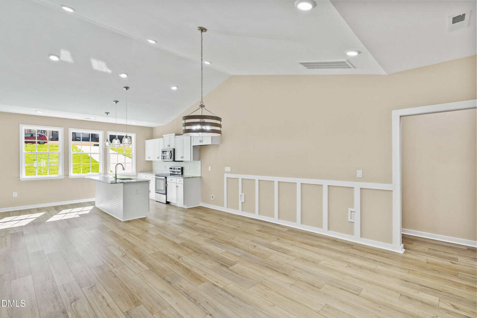 Bright open-concept kitchen and living area with light wood floors, beige walls, white cabinetry, pendant lighting, and large windows letting in natural light.