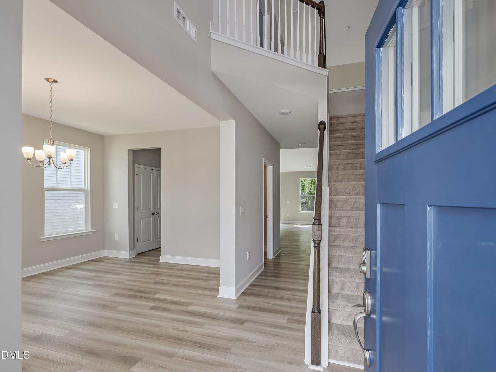 View from an open blue front door at 240 Sugar Pine Drive reveals a welcoming foyer with wood flooring, carpeted stairs featuring a white railing, a dining area adorned with a chandelier, and a hallway leading to other rooms. Enjoy bright natural light and crisp white walls throughout this inviting Franklinton home.