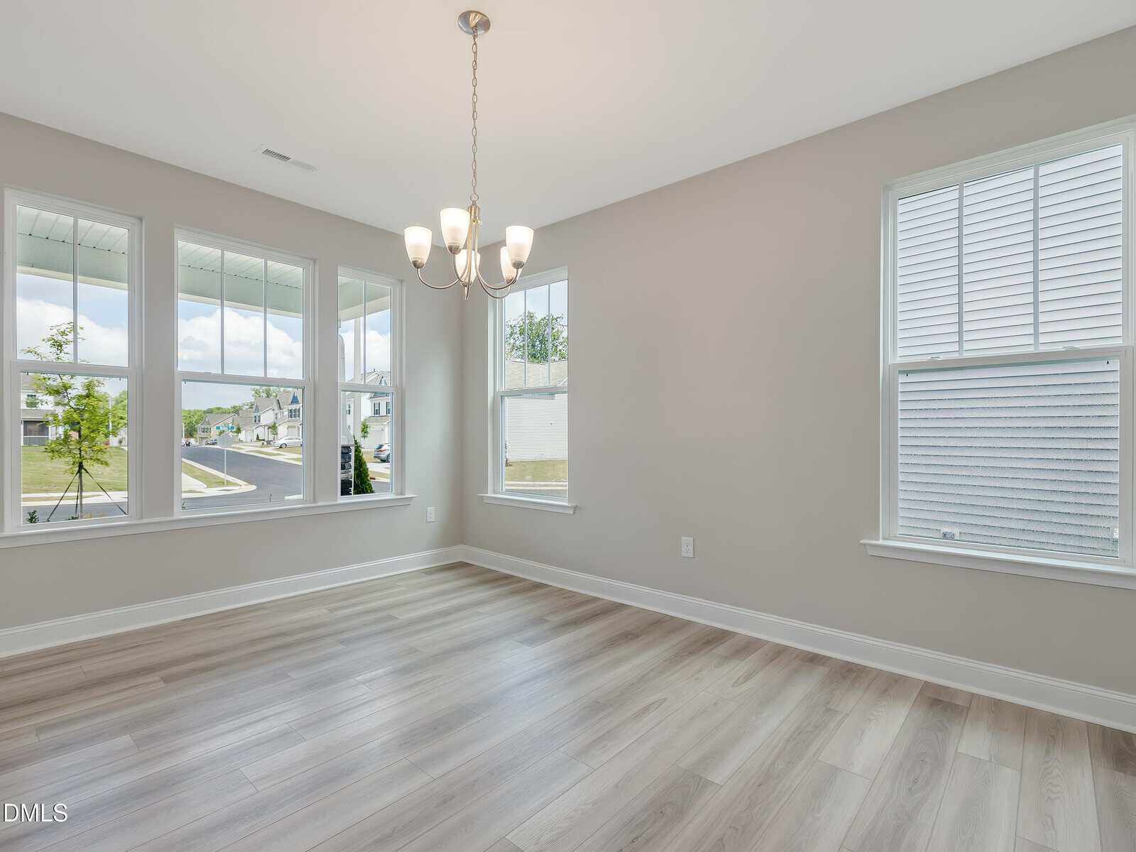 A bright, empty room with light-colored wood flooring, gray walls, three large windows, and a modern chandelier hanging from the ceiling. The windows overlook a street and neighboring house.