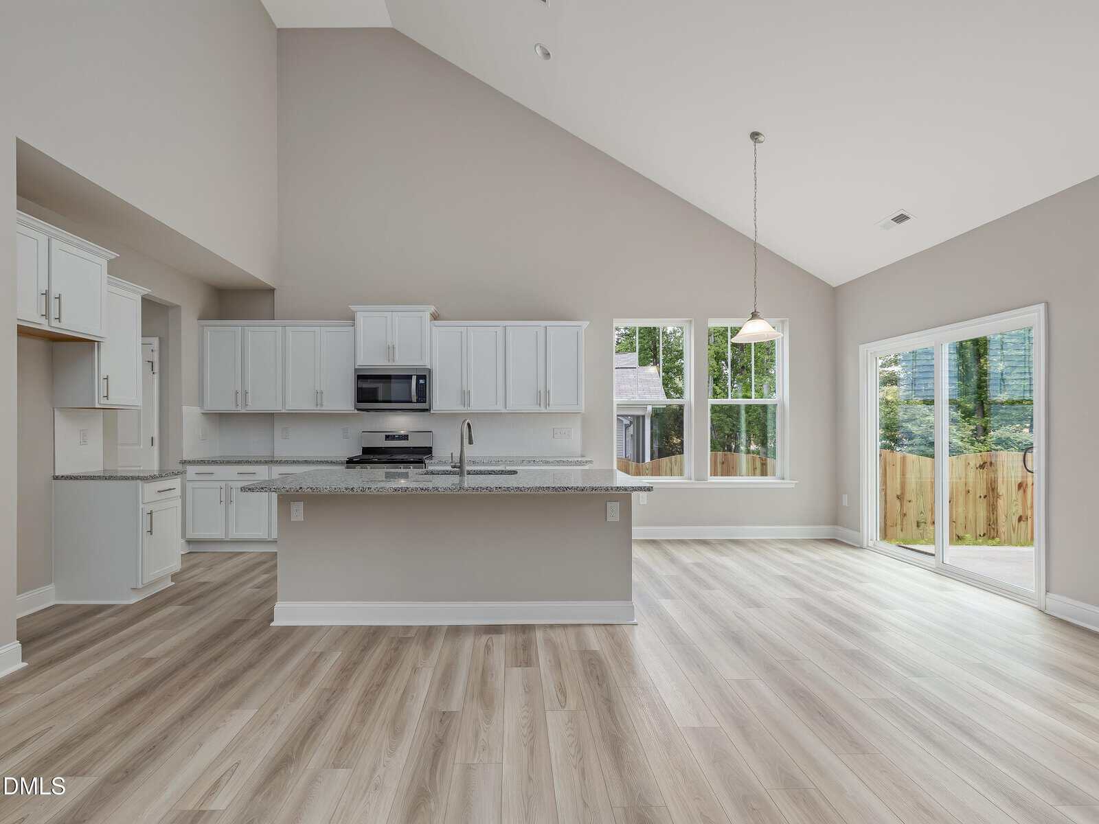 Modern kitchen and dining area with white cabinets, stainless steel appliances, an island, light wood flooring, large windows, and a sliding glass door leading to a fenced backyard. Neutral colors and high, sloped ceiling are visible.