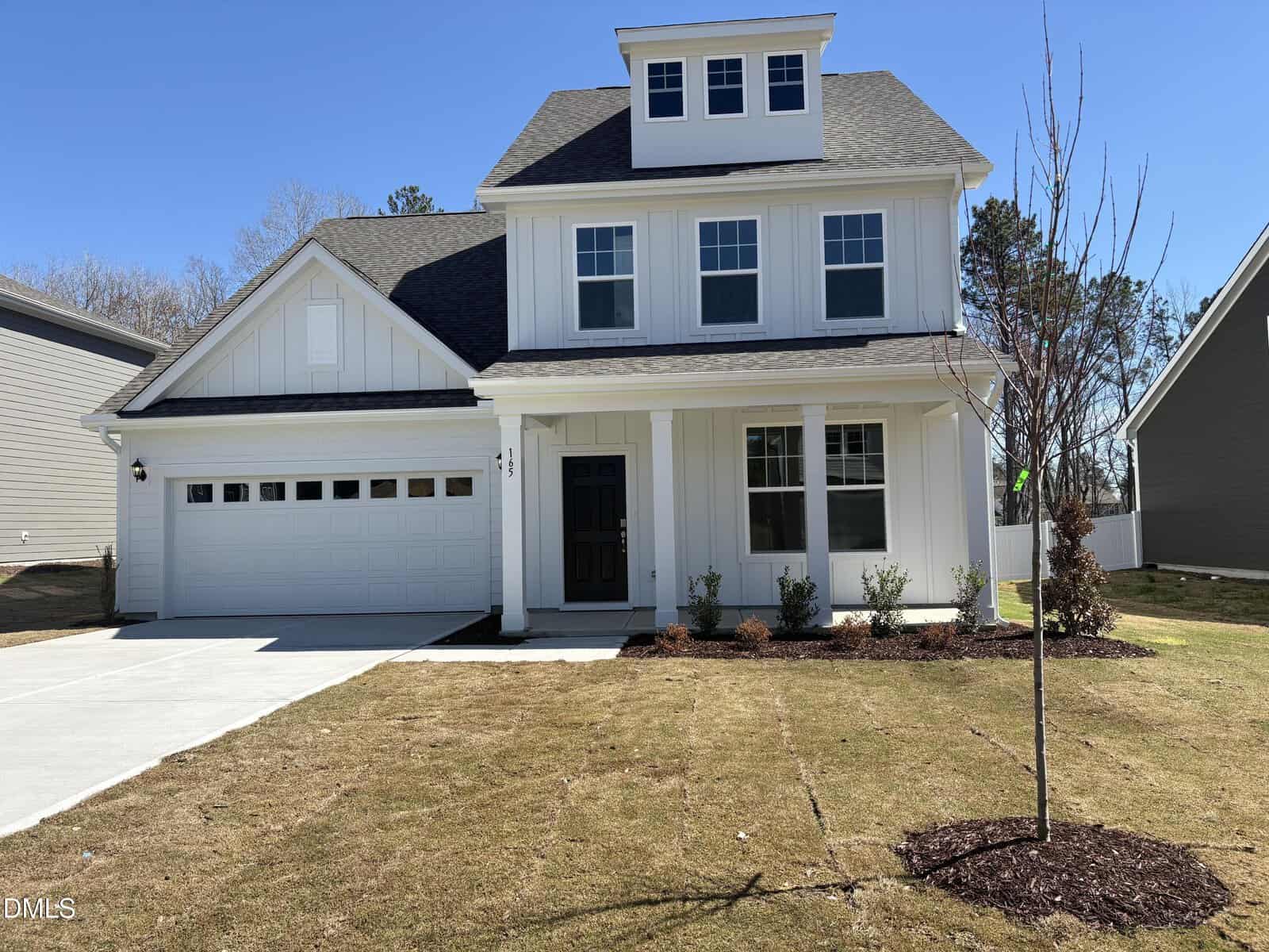 Two-story white house with a front porch, black door, attached two-car garage, and a small tree in the yard. The lawn is patchy, and the sky is clear and blue.