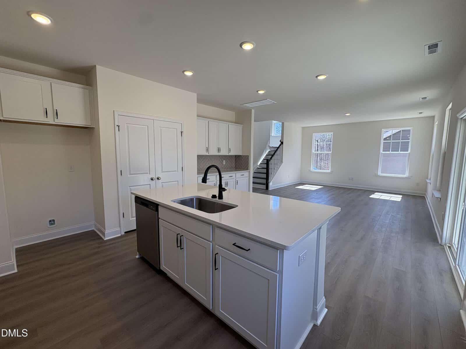 Spacious modern kitchen with white cabinets, island sink, stainless steel appliances, and open floor plan leading to a bright living area with large windows and wooden floors. Stairs are visible in the background.