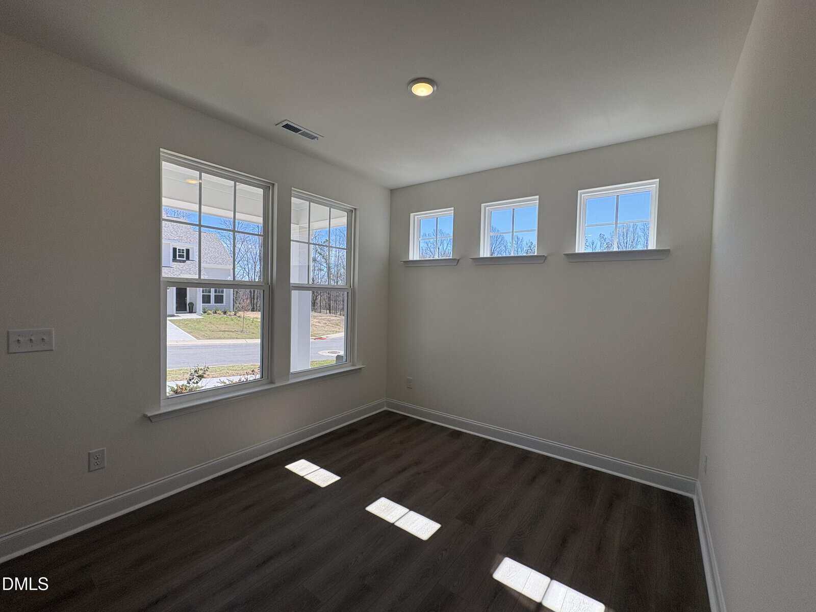 Bright room with large front-facing windows and three small upper windows, white walls, recessed ceiling light, and dark wood flooring; view of neighborhood houses and lawn outside.