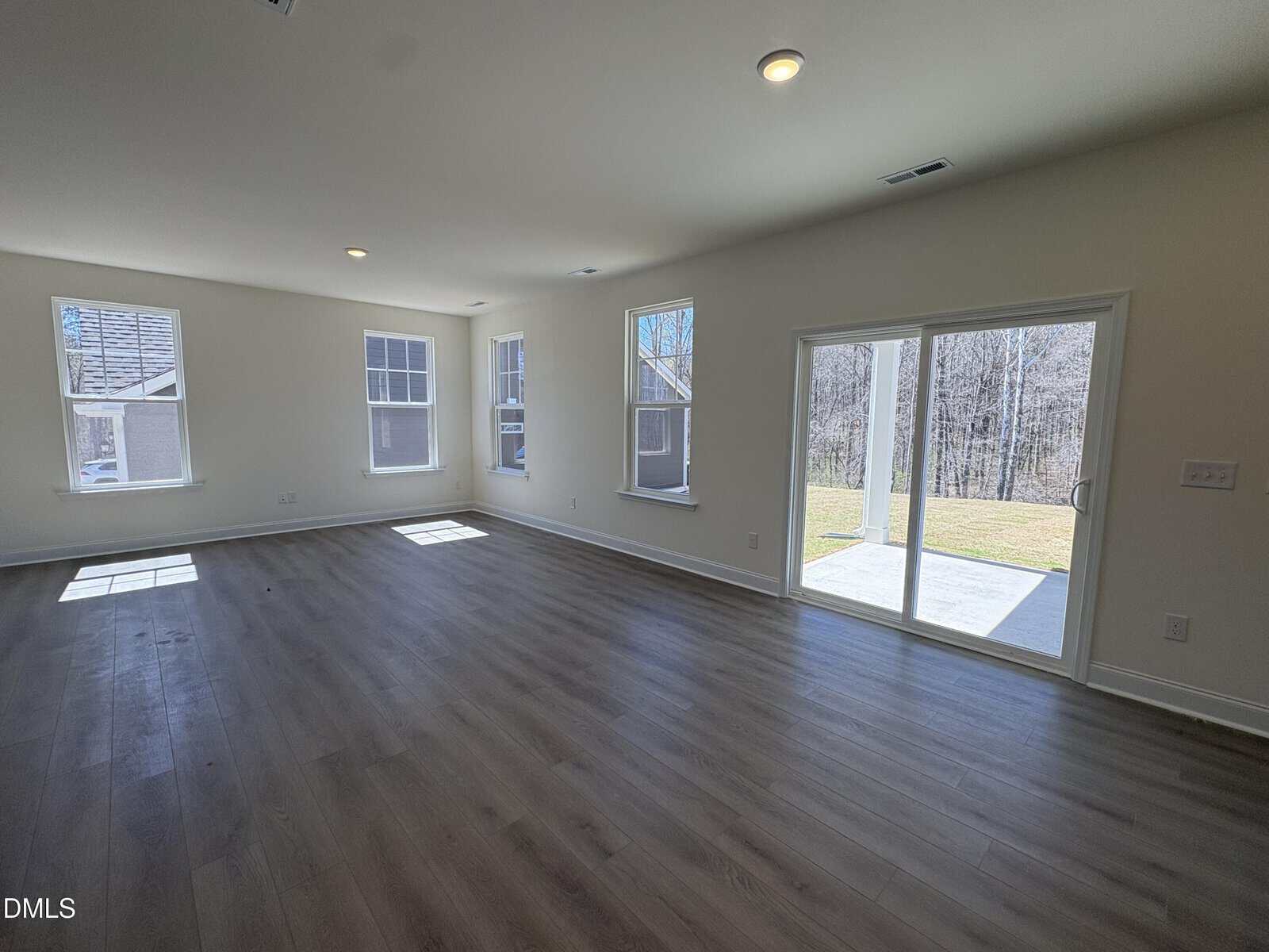 Bright, empty living room with large windows, wood flooring, and a sliding glass door opening to a patio with a view of trees and grass outside.