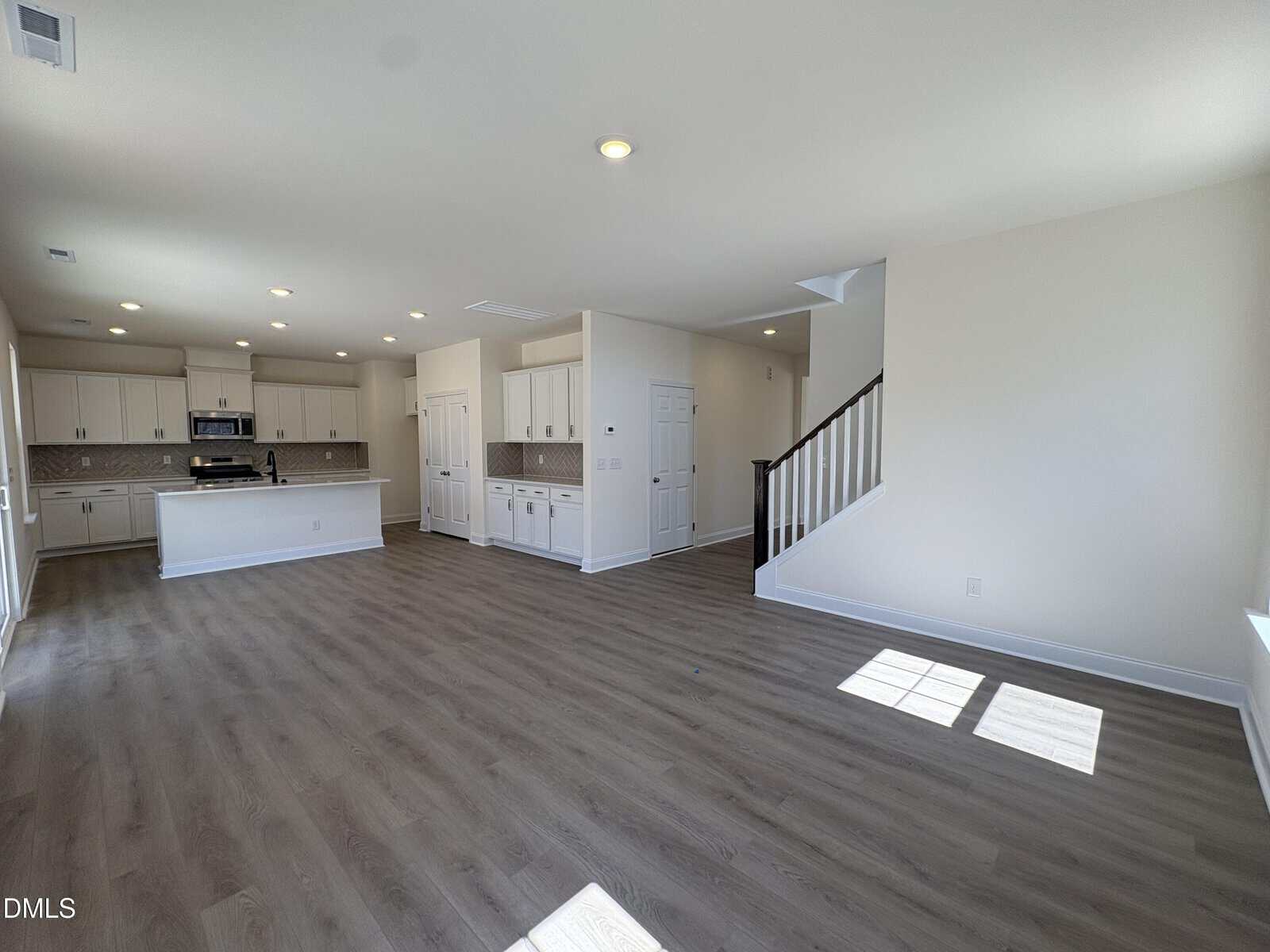 Spacious open-concept living area with wood flooring, white walls, modern kitchen, island, and staircase leading upstairs. Large windows let in natural light.