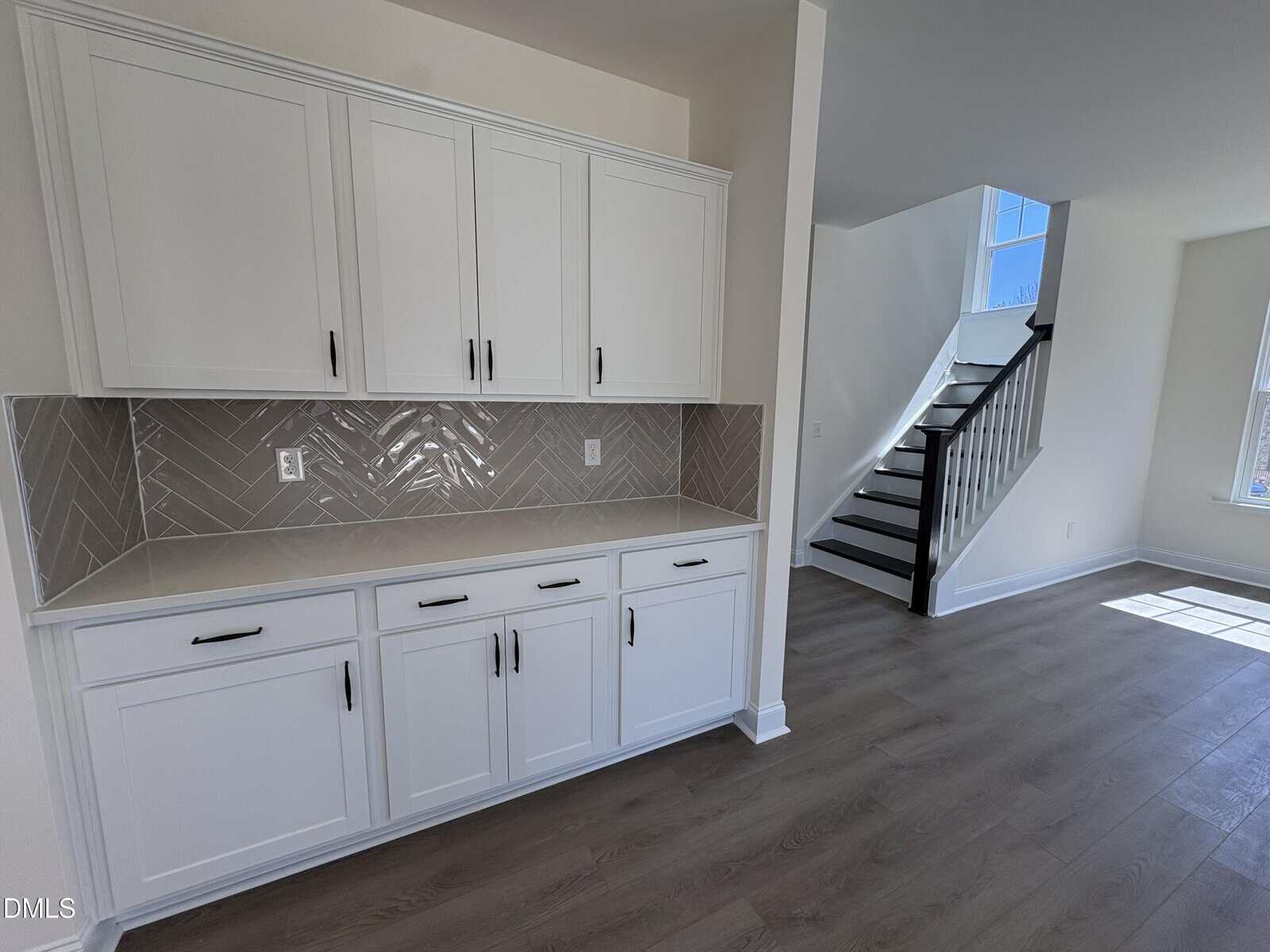 White kitchen cabinets with black handles, a beige herringbone tile backsplash, light wood floors, and a staircase with dark steps and white risers in a bright, modern home interior.