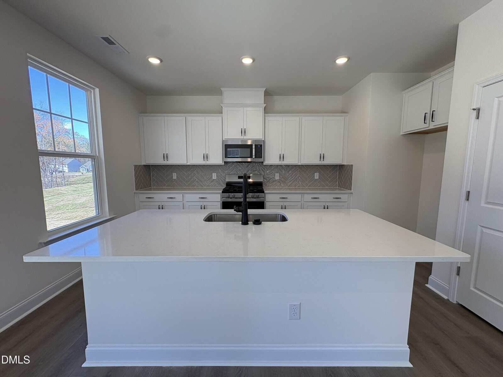 Modern kitchen with white cabinets, a large white island with a black faucet, built-in microwave, and patterned tile backsplash. Natural light enters through a window on the left, highlighting the neutral color scheme.