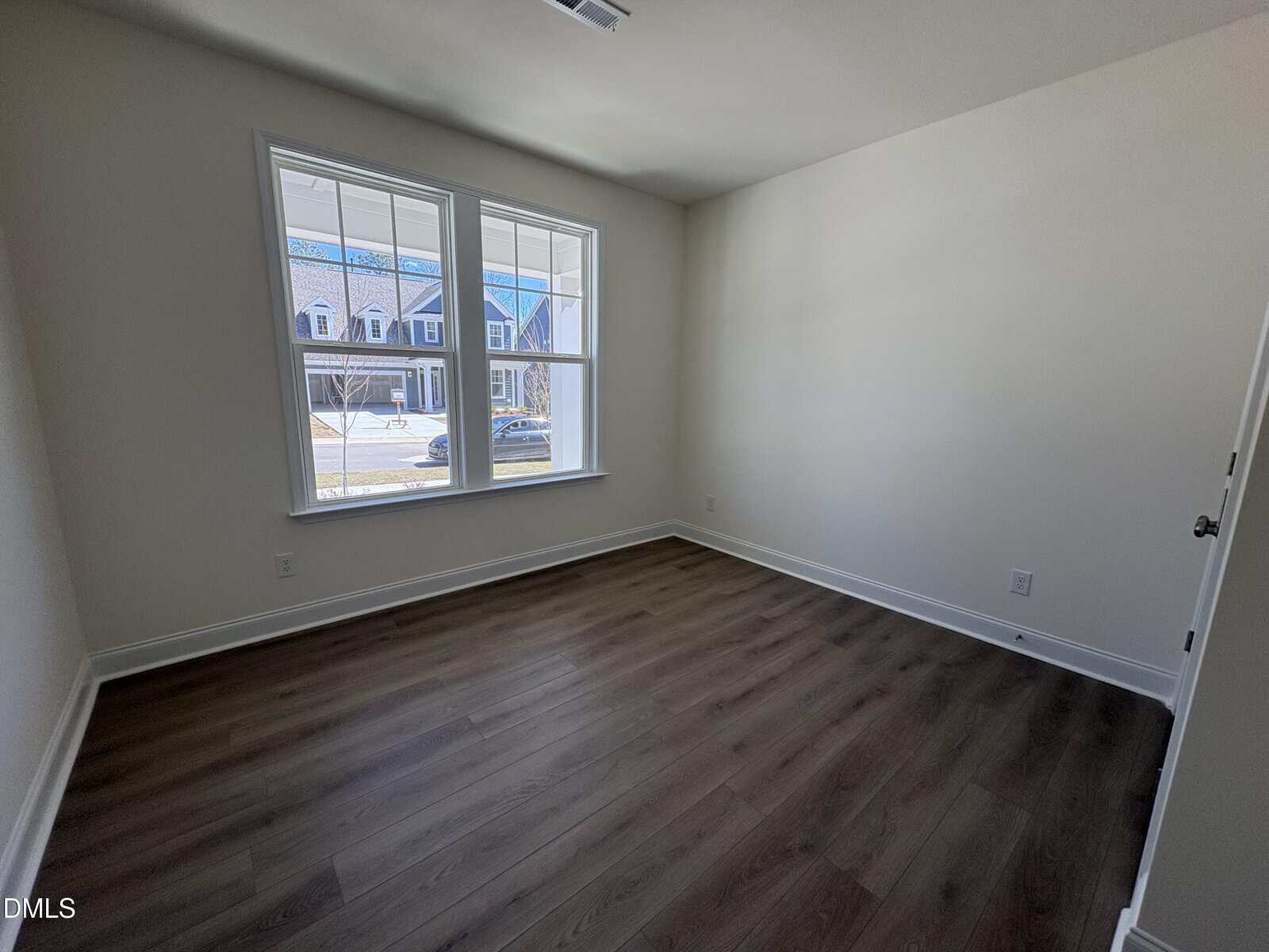 A bright, empty room with white walls, dark wood flooring, and a large window letting in natural light, showing houses and a car outside.