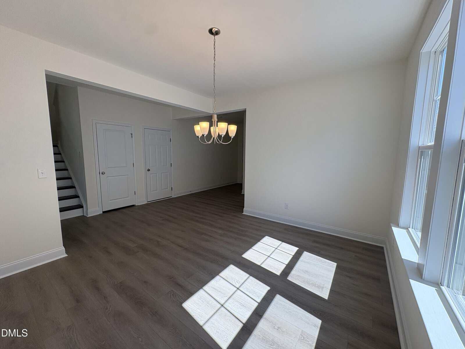 Bright, empty room with large windows, wood flooring, a modern chandelier, double doors, and a staircase in the background. Sunlight streams in, casting shadows on the floor.